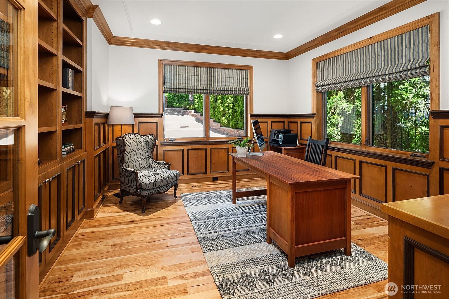 This sophisticated home office features rich wood paneling, built-in bookshelves, and warm hardwood flooring, creating a classic and professional atmosphere. A large wooden desk is positioned centrally, complemented by a patterned area rug and a comfortable patterned armchair near the window. The room is bathed in natural light from two large windows, offering a serene view of the outdoors.