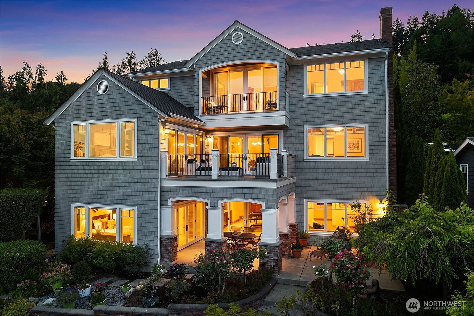 This stunning three-story residence features classic shingle-style architecture with multiple levels of outdoor living space, including a covered patio and two upper-level balconies. The warm, inviting glow from the interior lighting contrasts beautifully against the twilight sky, highlighting the home's elegant windows and well-manicured garden landscaping. The perspective captures the full rear facade, emphasizing the seamless transition between the indoor living areas and the lush, tiered backyard.