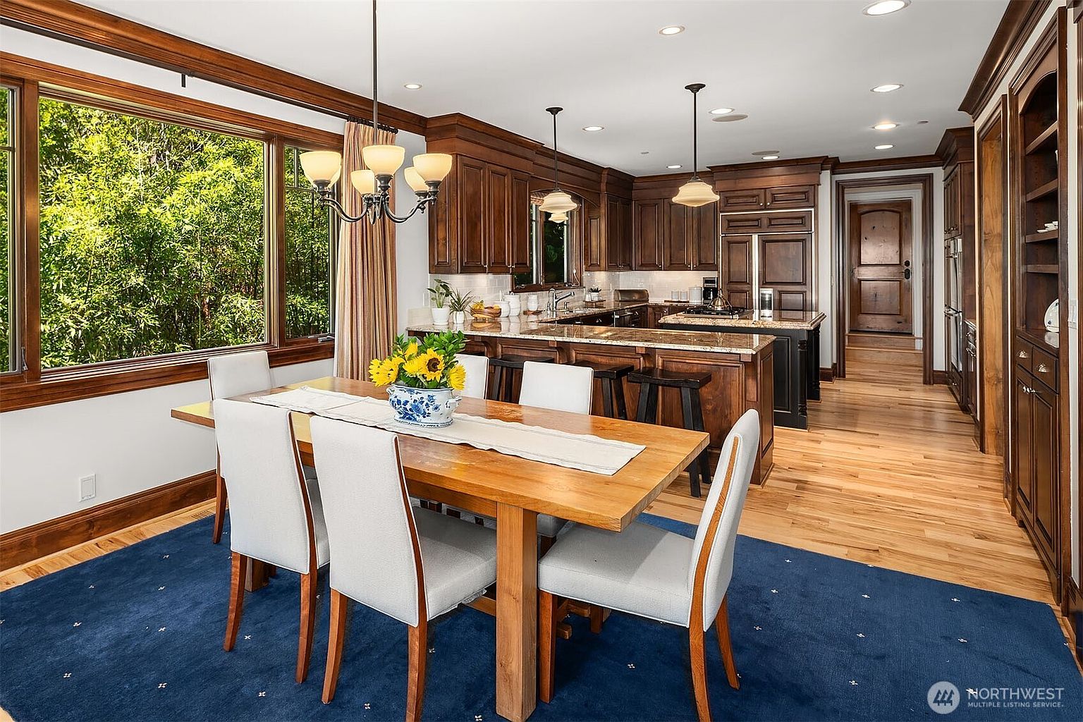 This elegant dining area features a warm wooden table set against a vibrant blue rug, seamlessly transitioning into an open-concept kitchen with rich, dark wood cabinetry and granite countertops. Large windows flood the space with natural light, highlighting the sophisticated blend of traditional woodwork and modern amenities. The perspective captures the inviting flow between the dining and culinary spaces, emphasizing a comfortable and high-end residential atmosphere.