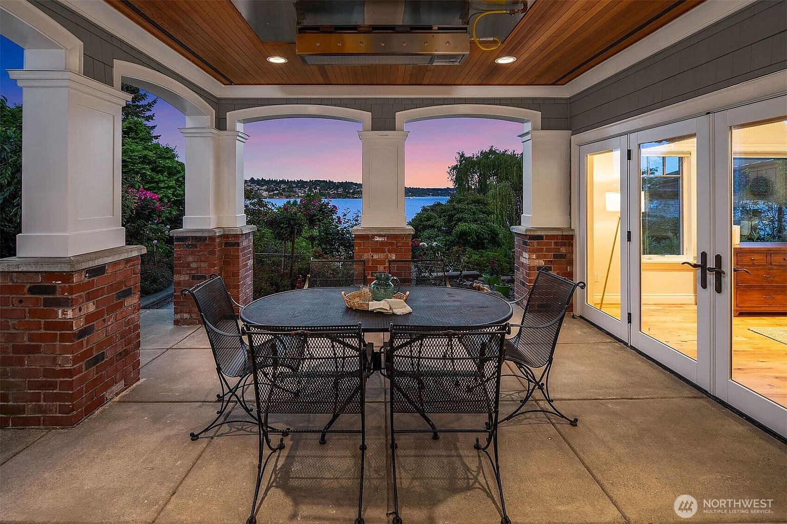 This covered patio offers a sophisticated outdoor dining space with a wrought-iron table and chairs set against a stunning sunset view of the water. The area features a warm wood-paneled ceiling with recessed lighting, brick-based columns, and large glass doors that seamlessly connect the indoor and outdoor living spaces. The overall atmosphere is serene and luxurious, perfect for evening relaxation or entertaining.