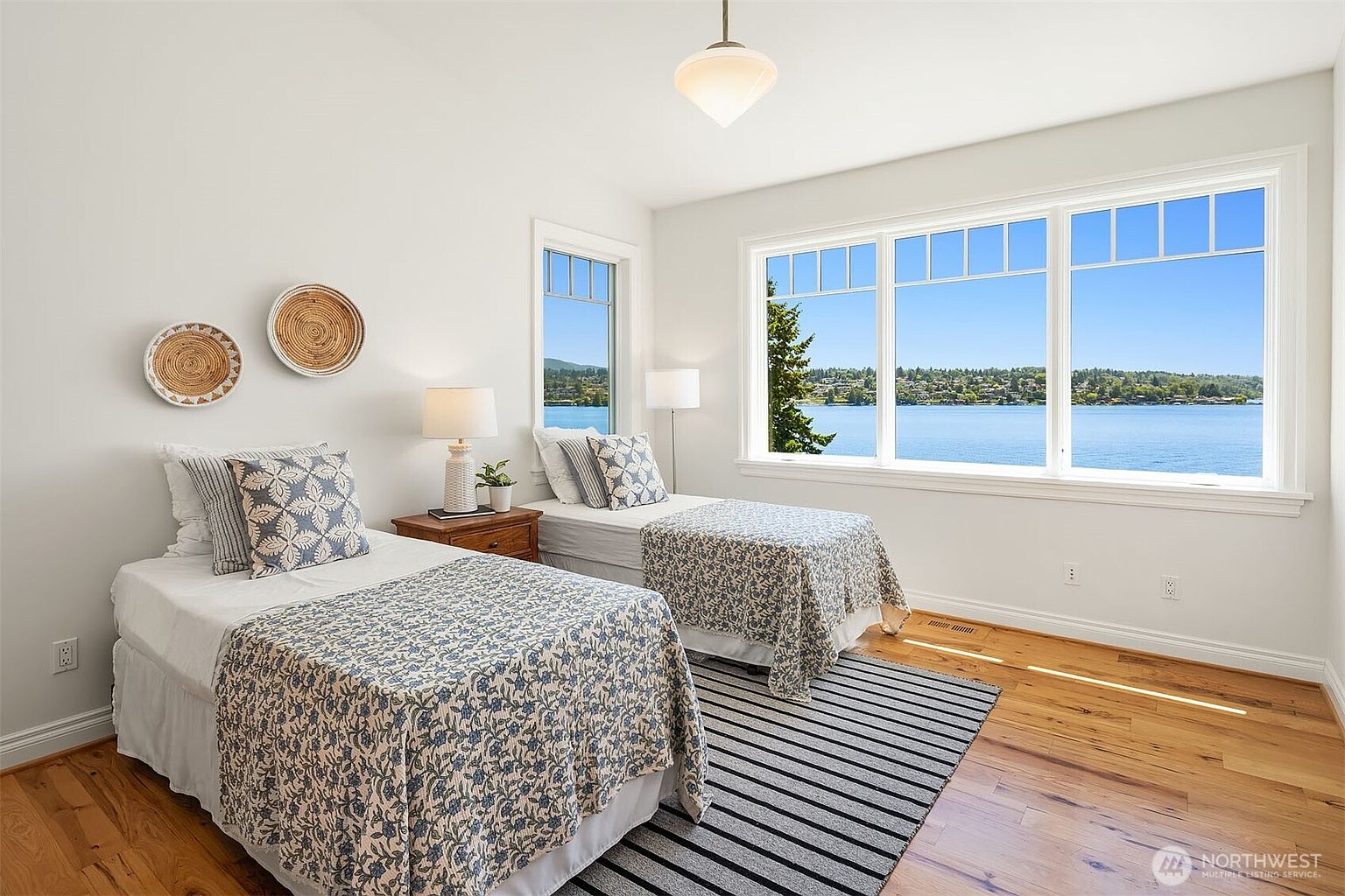 This bright and airy guest bedroom features two twin beds with matching blue and white floral patterned quilts, positioned to take full advantage of the expansive water views through large windows. The room is decorated with a coastal-inspired aesthetic, including woven wall art and a striped area rug, creating a serene and inviting atmosphere. The warm hardwood floors and neutral walls enhance the natural light, making the space feel open and tranquil.