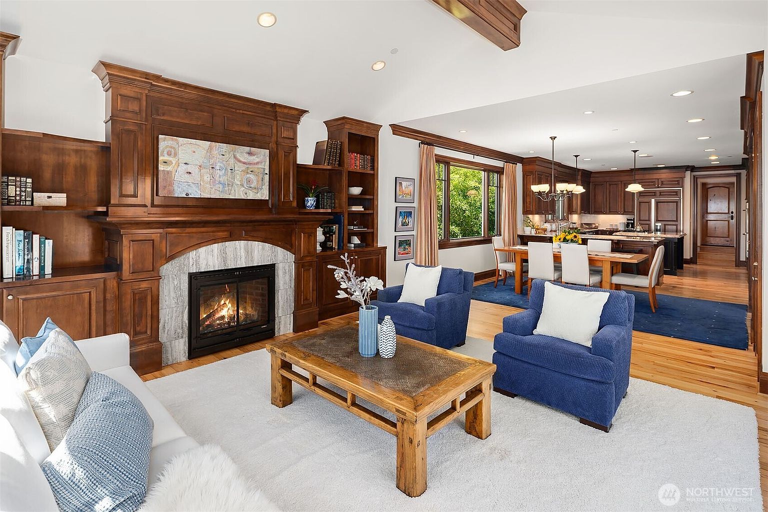 This spacious, open-concept living room features a grand wood-paneled fireplace as its focal point, complemented by built-in shelving and a cozy seating area with plush blue armchairs and a white sofa. The room flows seamlessly into a formal dining area and kitchen, creating an inviting and cohesive living space with warm hardwood floors and elegant architectural details. The perspective is a wide-angle shot from the living area looking toward the dining space, highlighting the home's airy and well-appointed layout.