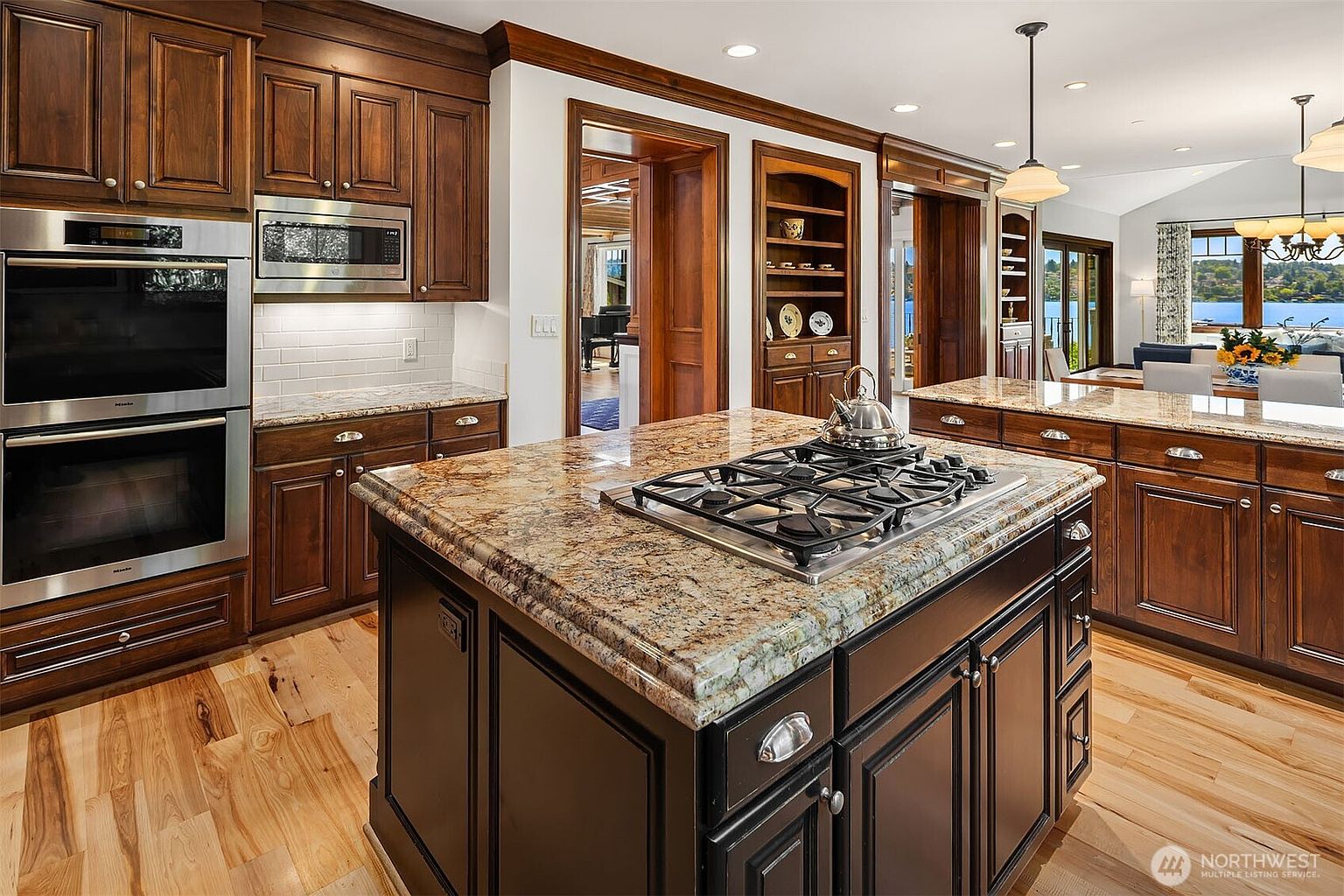 This elegant kitchen features rich, dark-stained wooden cabinetry paired with warm, granite countertops and a central island equipped with a gas cooktop. The space is anchored by high-end stainless steel appliances, including a double wall oven, and flows seamlessly into an adjacent dining area with a scenic water view. The combination of natural hardwood flooring and traditional wood trim creates a sophisticated, warm, and inviting atmosphere.