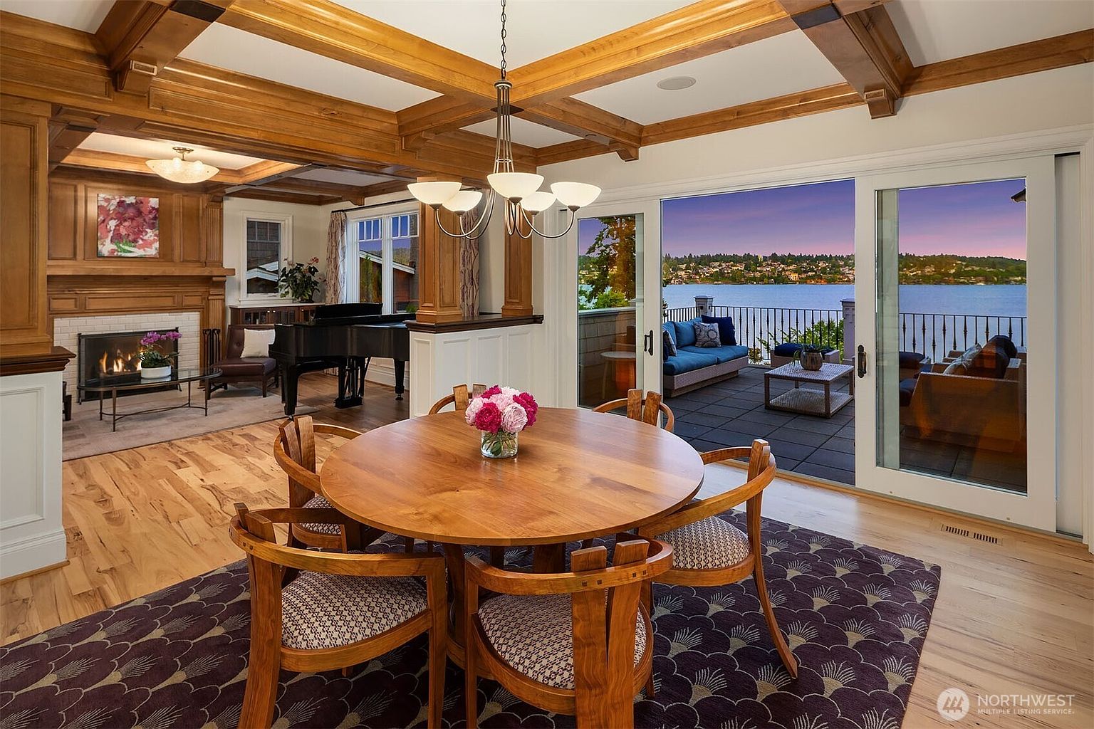 This elegant dining area features a warm, circular wooden table as its centerpiece, set beneath a sophisticated chandelier with a coffered wood-beam ceiling above. The room flows seamlessly into a living space with a fireplace and piano, while large sliding glass doors open to a scenic deck overlooking a tranquil lake at sunset. The combination of rich wood tones, classic architectural details, and expansive water views creates a luxurious and inviting atmosphere.