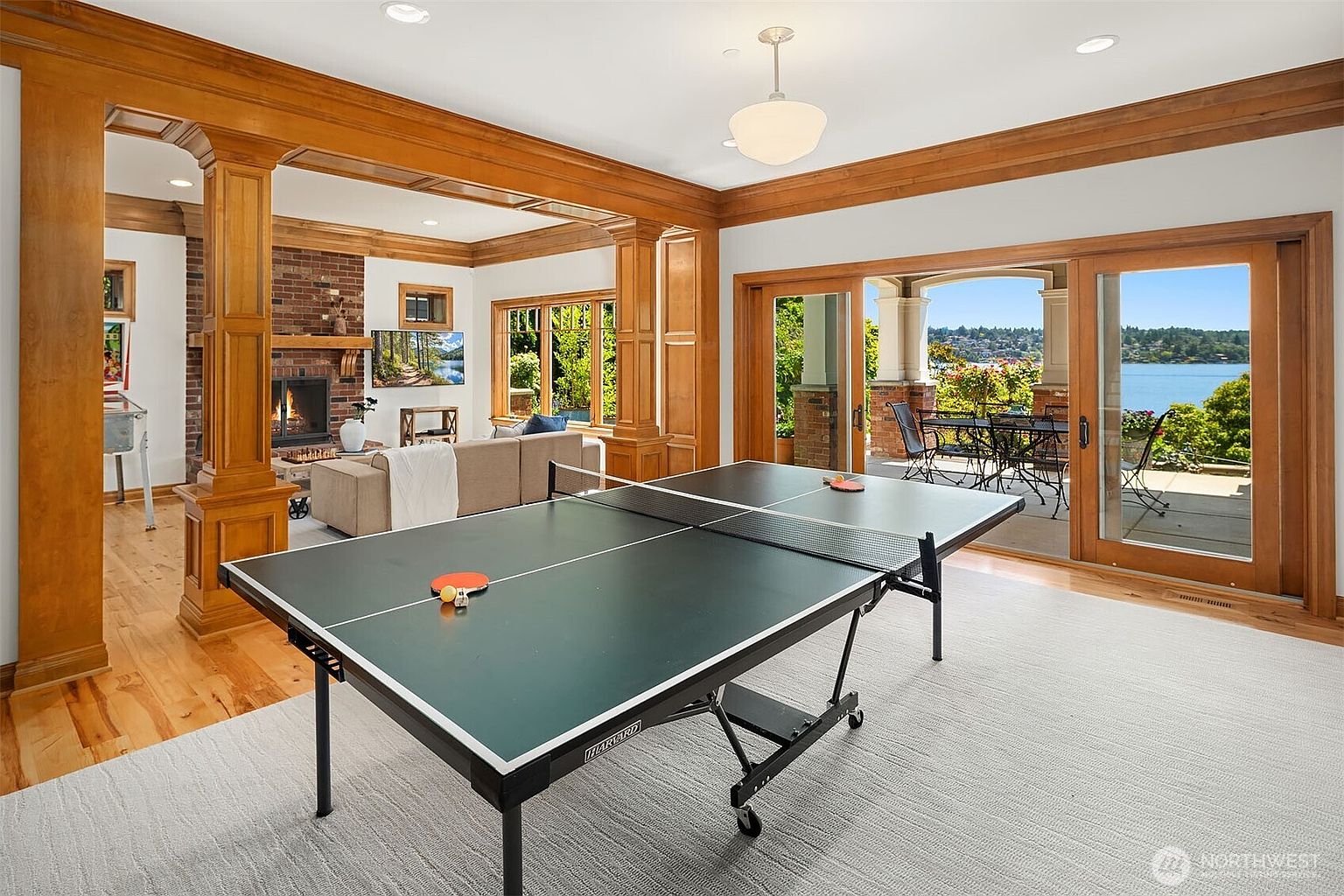 This spacious, open-concept living area features a prominent ping pong table in the foreground, transitioning into a cozy seating area with a brick fireplace and a wall-mounted television. The room is framed by rich, warm-toned wood trim and columns, with large sliding glass doors that open onto a scenic patio overlooking a tranquil lake. The overall aesthetic is a blend of comfortable, traditional craftsmanship and inviting, light-filled leisure space.