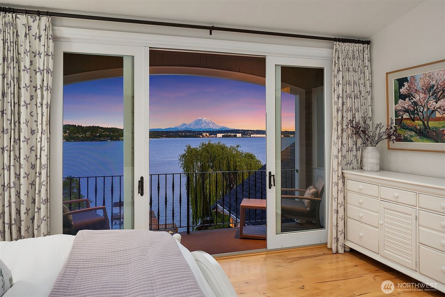 This elegant primary bedroom features a stunning, unobstructed view of a lake and a snow-capped mountain through large sliding glass doors that open onto a private balcony. The room is decorated with soft, neutral tones, including patterned curtains and a classic white dresser, creating a serene and luxurious atmosphere. The warm hardwood flooring and the picturesque sunset backdrop emphasize the home's premium waterfront location.