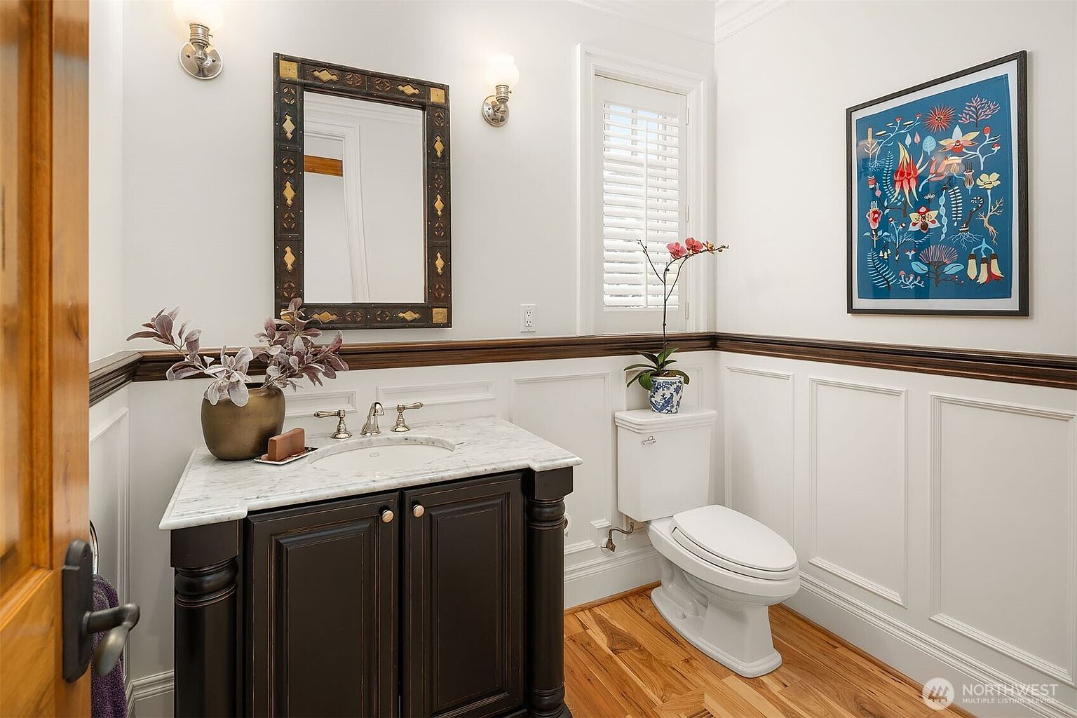 This elegant powder room features a dark wood vanity topped with a white marble countertop and a decorative framed mirror. The walls are adorned with classic white wainscoting and a vibrant botanical art piece, creating a sophisticated and welcoming atmosphere. The space is completed by a white toilet, hardwood flooring, and a window with plantation shutters that allows for soft, natural light.