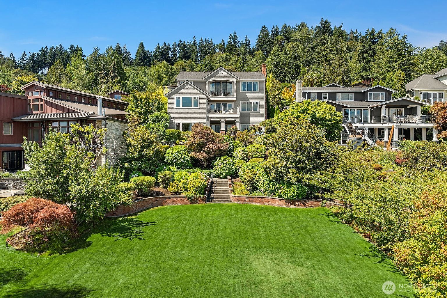 This elevated rear view captures a sprawling, meticulously manicured lawn leading up to a tiered landscape of lush greenery and mature trees. The perspective highlights the multi-level architecture of the homes, showcasing expansive decks and large windows that overlook the serene, private backyard setting. The scene conveys a sense of luxury and tranquility, emphasizing the seamless integration between the residential structures and the natural, sloped terrain.