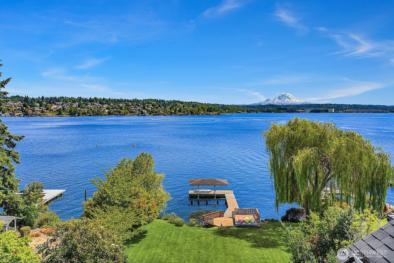 This elevated rear view captures a stunning waterfront property overlooking a vast lake with a prominent snow-capped mountain in the distance. The scene features a lush green lawn leading down to a private wooden dock with a covered boat lift and a small deck area with colorful seating. The composition emphasizes the serene, expansive landscape and the direct water access, highlighting the property's premium location.