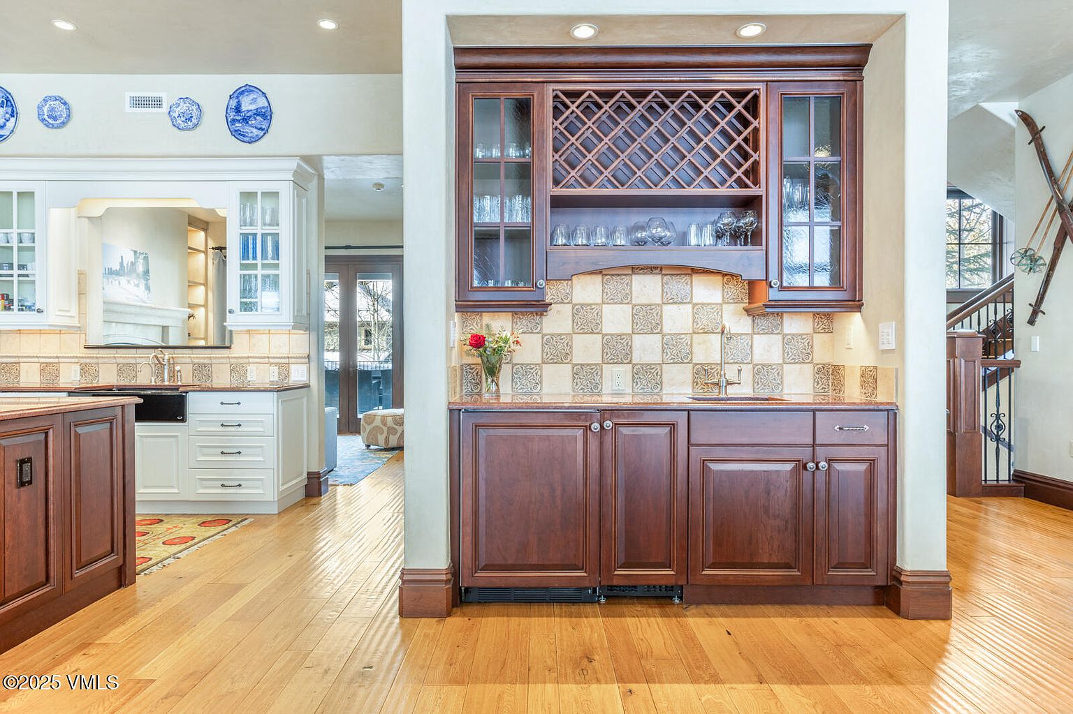 This interior shot showcases a well-appointed kitchen with a mix of dark wood and white cabinetry. The dark wood bar area features a wine rack and decorative tile backsplash, while the adjacent kitchen area has white cabinets and a contrasting countertop. The hardwood flooring adds warmth to the space, and the overall impression is one of classic elegance and functionality.