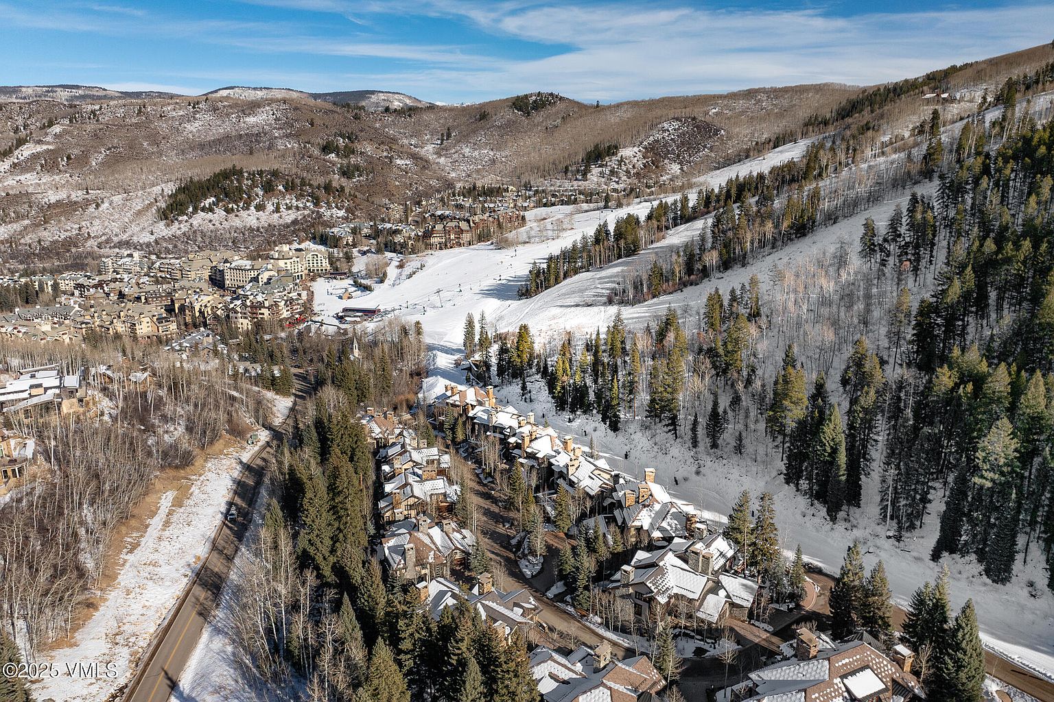 This aerial view showcases a luxurious mountain resort community nestled amidst snow-covered slopes and evergreen forests. The architecture features a mix of chalet-style homes with steeply pitched roofs, blending seamlessly with the natural landscape. A winding road leads through the community, offering access to the ski slopes and nearby town, creating an inviting and picturesque setting.
