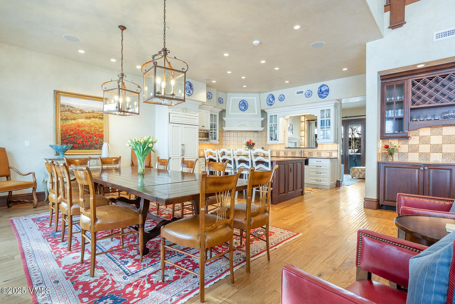 This interior shot showcases a dining room seamlessly connected to a kitchen area. The dining room features a large wooden table surrounded by chairs, set upon a patterned rug, with two lantern-style chandeliers hanging above. The kitchen boasts white cabinetry, a dark wood island, and decorative blue plates adorning the upper walls, creating a warm and inviting atmosphere.