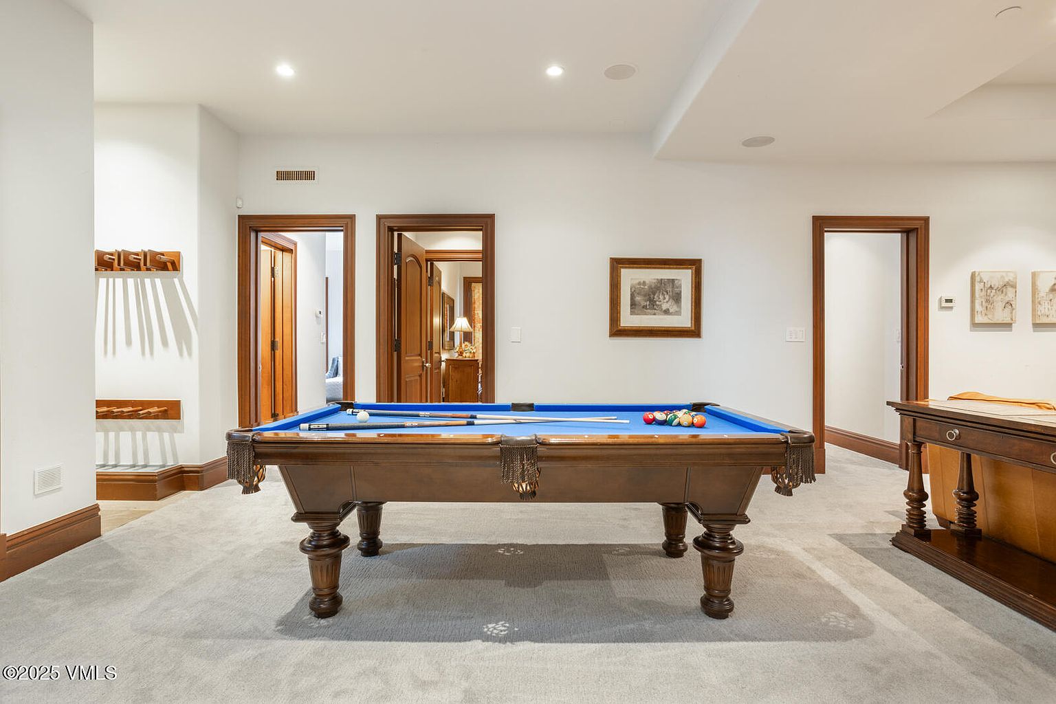 This interior shot showcases a basement recreation area featuring a classic wooden pool table with blue felt, complete with cues and billiard balls. The room is well-lit with recessed lighting and features neutral-toned walls, complemented by dark wood trim around the doorways and baseboards. The perspective is centered on the pool table, emphasizing the room's entertainment focus.