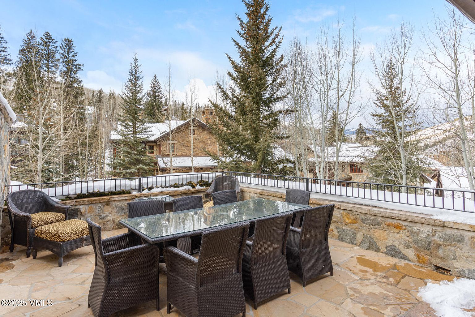 This image showcases a spacious patio or deck area, furnished with a large outdoor dining table and chairs, along with a comfortable lounge chair and ottoman. The patio is constructed with stone and features a low stone wall with a black metal railing, offering views of snow-covered trees and houses in the background. The scene evokes a sense of luxury and relaxation in a mountain setting.