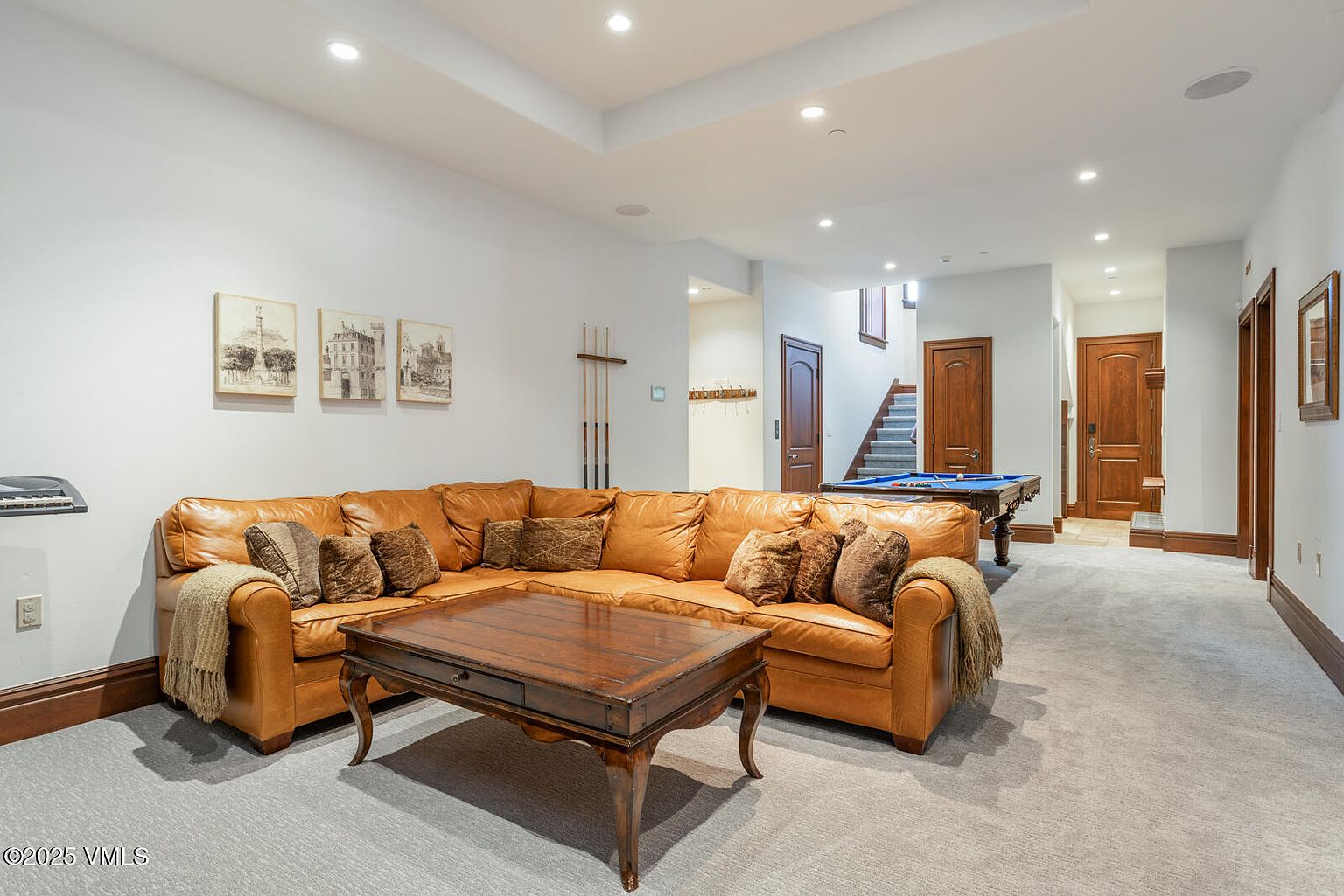 This is a well-appointed basement featuring a comfortable leather sectional sofa, a wooden coffee table, and a pool table. The room is illuminated by recessed lighting and includes artwork on the walls. The space is carpeted and has wood trim, creating a cozy and inviting atmosphere for relaxation and entertainment.