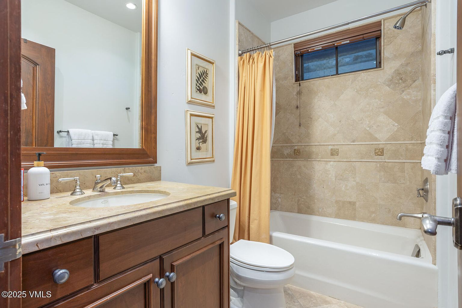 This is a well-lit bathroom featuring a wooden vanity with a light-colored countertop and a sink. Above the vanity is a large mirror with a wooden frame. To the right, there is a bathtub with a shower, enclosed by a beige tile surround and a yellow shower curtain. A toilet is positioned next to the tub, and framed botanical prints adorn the wall above it.