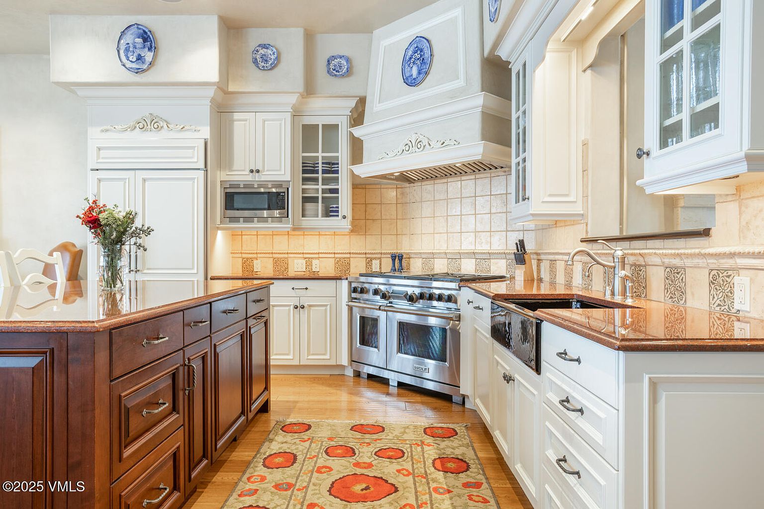 This is a well-appointed kitchen featuring custom white cabinetry, a stainless steel Wolf range, and a contrasting dark wood island with a polished countertop. The backsplash is tiled, and decorative blue and white plates adorn the walls above the cabinets. The perspective is from a medium distance, showcasing the layout and high-end finishes.