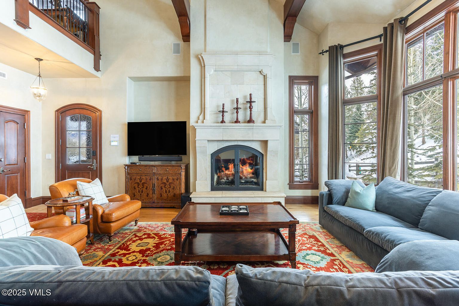 This is an interior shot of a luxurious living room featuring a stone fireplace as a focal point, flanked by a television and large windows offering a snowy outdoor view. The room is furnished with comfortable sofas, leather armchairs, and a patterned rug, creating a warm and inviting atmosphere. Exposed wooden beams add a rustic touch to the elegant space.