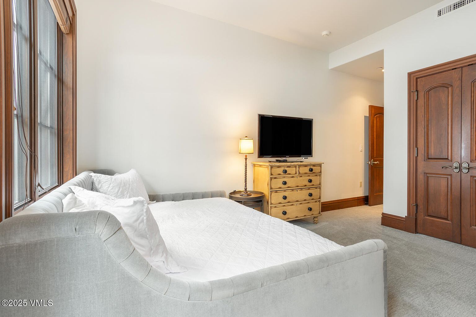 This is a cozy guest bedroom featuring a daybed with white bedding and pillows, positioned near a window with wooden trim. A wooden dresser with a television on top sits against the wall, illuminated by a table lamp. The room has a neutral color palette and is carpeted, creating a comfortable and inviting space.