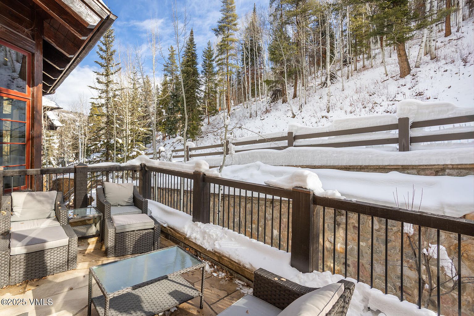 This image showcases a cozy patio or balcony area covered in snow, furnished with wicker seating and glass-topped tables. The space is bordered by a dark wood and iron railing, with a stone wall beneath, offering a view of a snow-covered hillside dotted with trees. The scene evokes a sense of winter tranquility and outdoor relaxation.