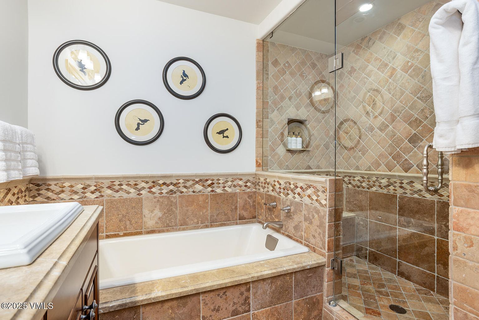 This is a well-appointed primary bathroom featuring a built-in bathtub with travertine tile surround and a glass-enclosed shower. The walls are adorned with framed artwork, and the space is illuminated by recessed lighting. The overall impression is one of luxury and relaxation.