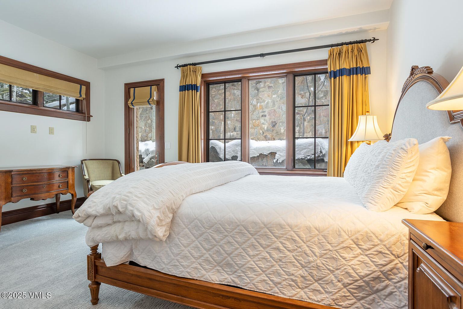 This is a cozy primary bedroom featuring a wooden bed frame with a white quilted comforter and pillows. The room has a window with gold and blue curtains, offering a view of a snowy landscape. A wooden dresser and a chair are visible on the left, adding to the room's warm and inviting atmosphere.