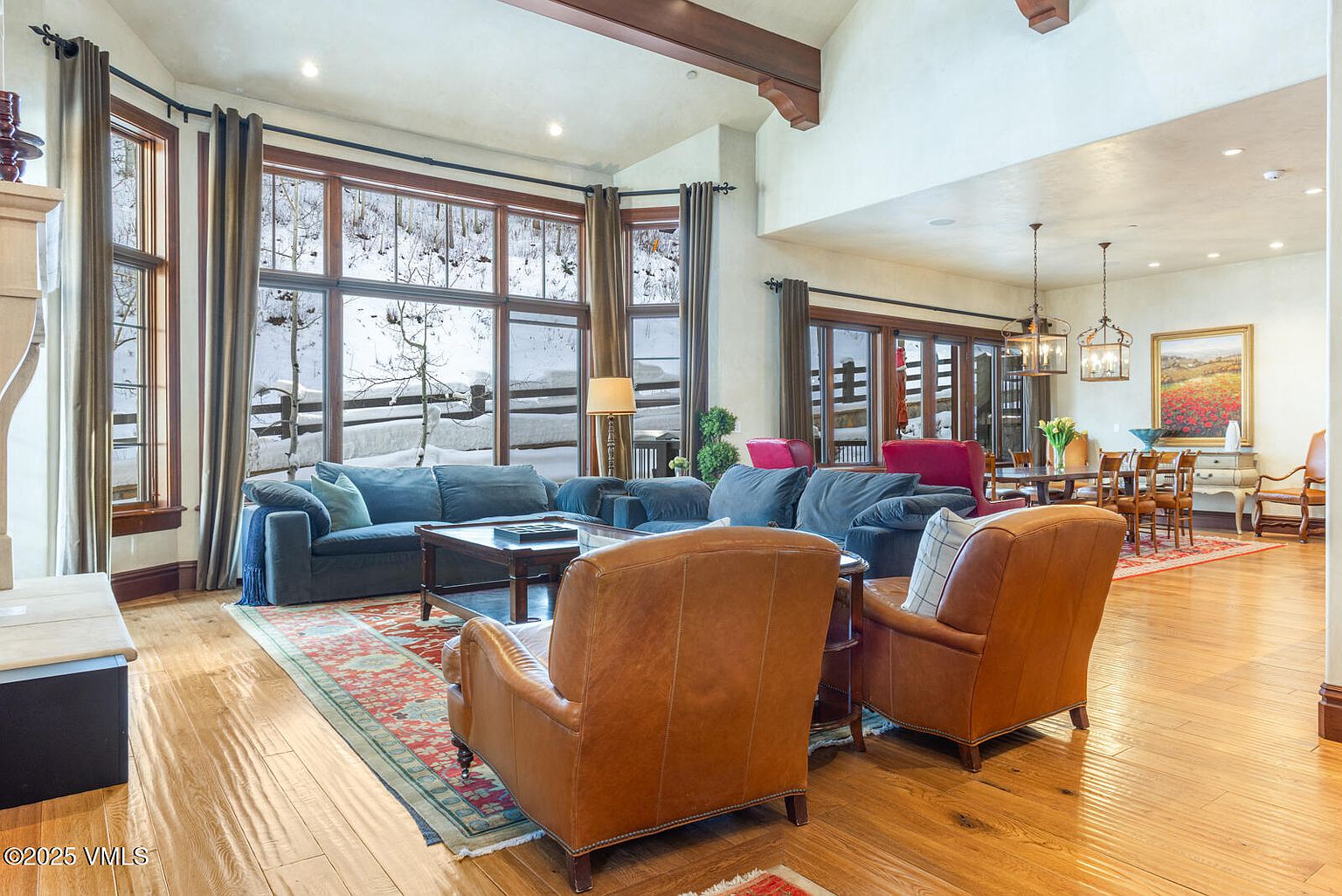 This is a spacious living room featuring hardwood floors, large windows with a snowy view, and comfortable seating arrangements. The room includes two blue sofas, two leather armchairs, and a coffee table, all arranged on a decorative rug. The high ceilings with wooden beams add a touch of rustic elegance to the space.