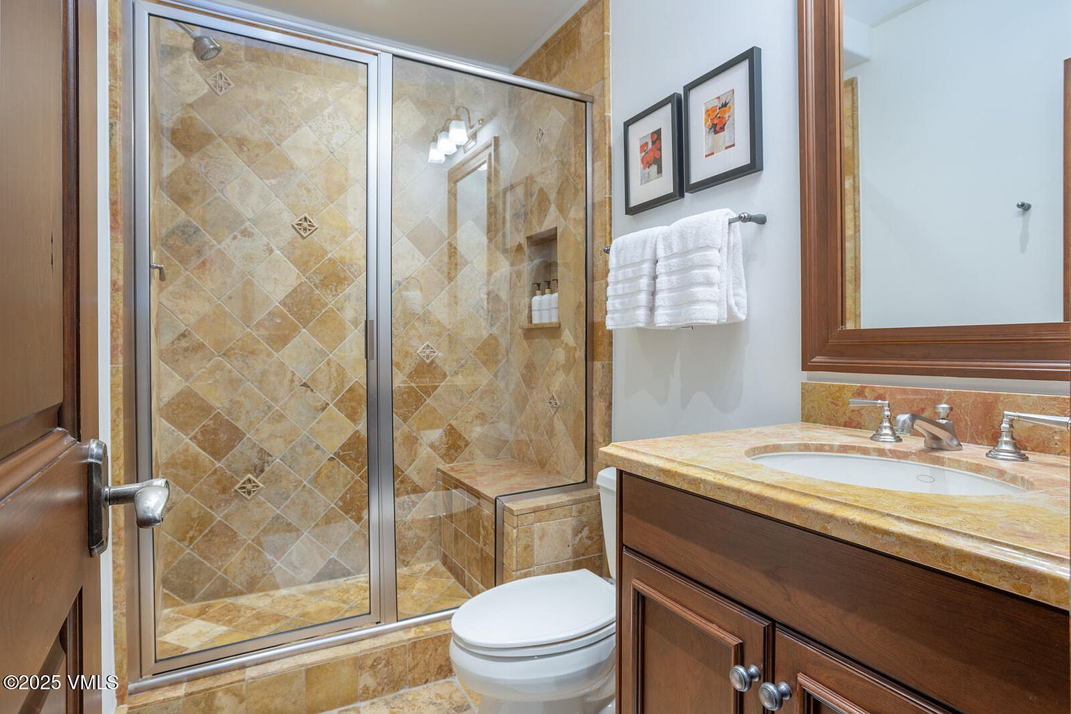 This is a well-appointed guest bathroom featuring a tiled shower with a glass door, a toilet, and a vanity with a marble countertop. The color palette is warm and inviting, with earth-toned tiles and dark wood cabinetry. Framed artwork and neatly arranged towels add a touch of elegance.