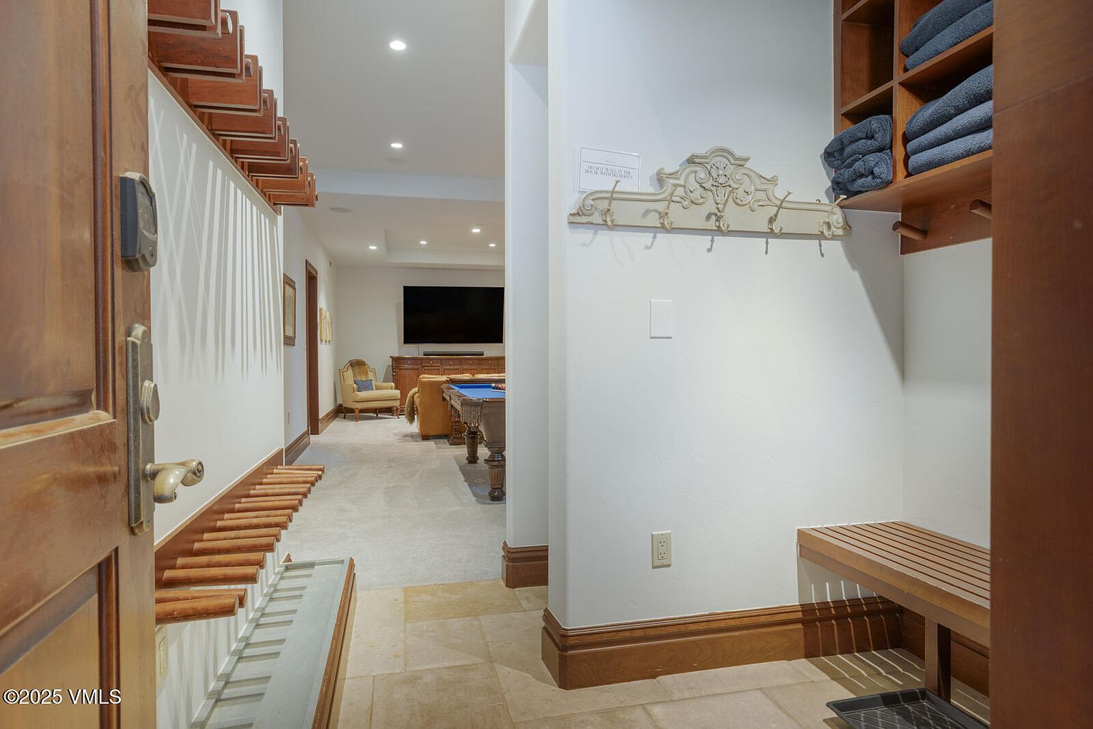 The image showcases an interior hallway leading to a recreational area, possibly a basement. The hallway features a unique design with wooden accents along the wall and ceiling, complemented by neutral-toned walls and tiled flooring. A glimpse into the recreational area reveals a pool table and seating, suggesting a space for entertainment and relaxation.