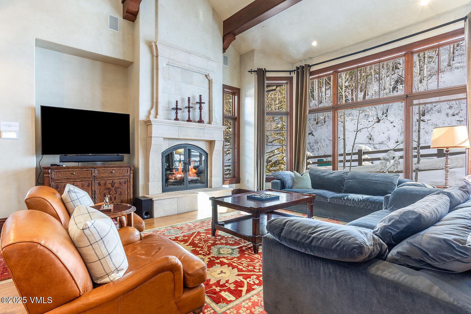 This is a warm and inviting living room featuring a stone fireplace, a large window with a view of a snowy landscape, and comfortable seating. The room is decorated with a mix of traditional and rustic elements, including exposed wooden beams and a patterned area rug. The overall impression is one of comfort and luxury.