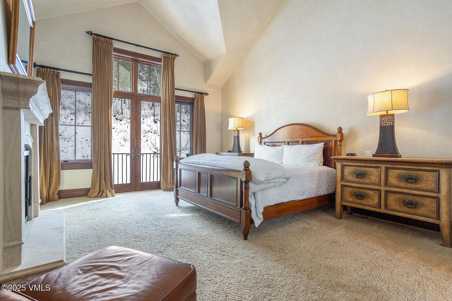 This is a primary bedroom featuring a wooden bed frame with white linens, a matching nightstand with a lamp, and a fireplace. The room has a neutral color palette with beige walls and carpet, and large windows with brown curtains provide natural light and a view of a snowy landscape. The overall impression is cozy and inviting.