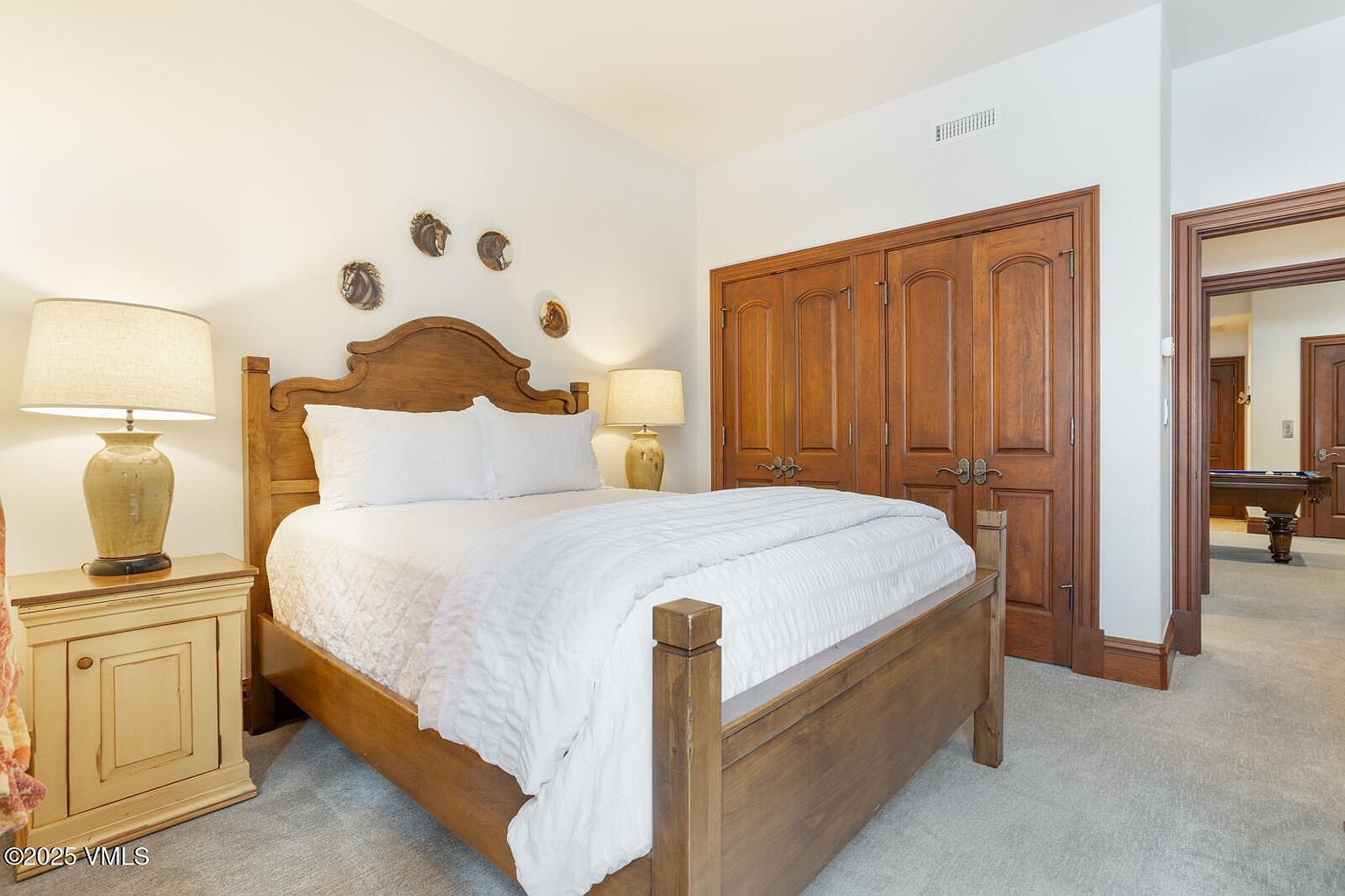 This is a cozy guest bedroom featuring a wooden bed frame with a white comforter and pillows. The room is decorated with horse-themed wall art above the bed. A wooden nightstand with a lamp sits beside the bed, and a large wooden closet is visible in the background, adding to the room's warmth and character.