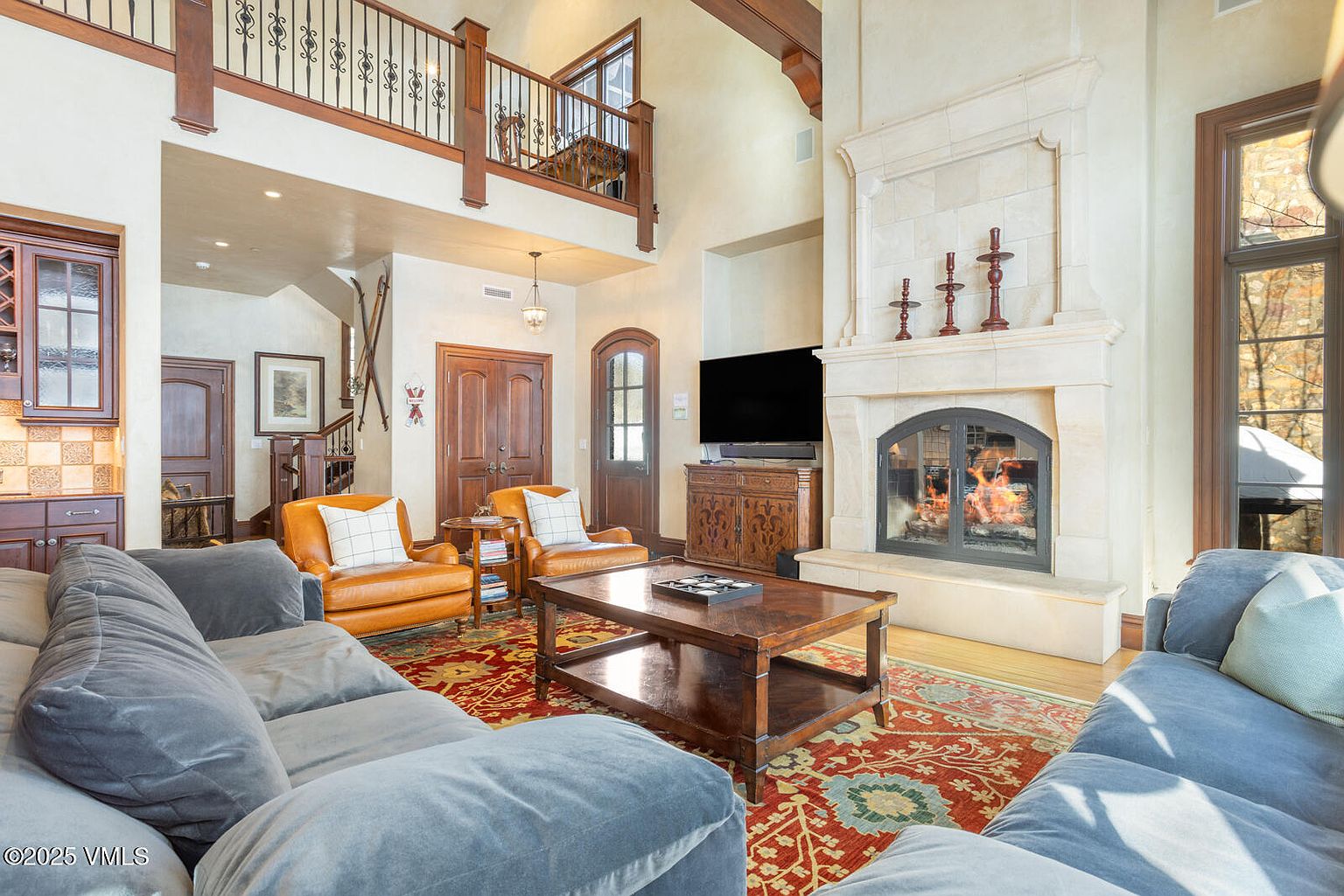 This is an interior shot of a luxurious living room featuring a stone fireplace with a burning fire, a large flat-screen TV, and comfortable seating including a gray sectional sofa and two leather armchairs. The room is decorated with a patterned rug and a wooden coffee table, and the high ceilings and architectural details create a grand and inviting atmosphere. The perspective is from the front of the room, capturing the depth and breadth of the space.