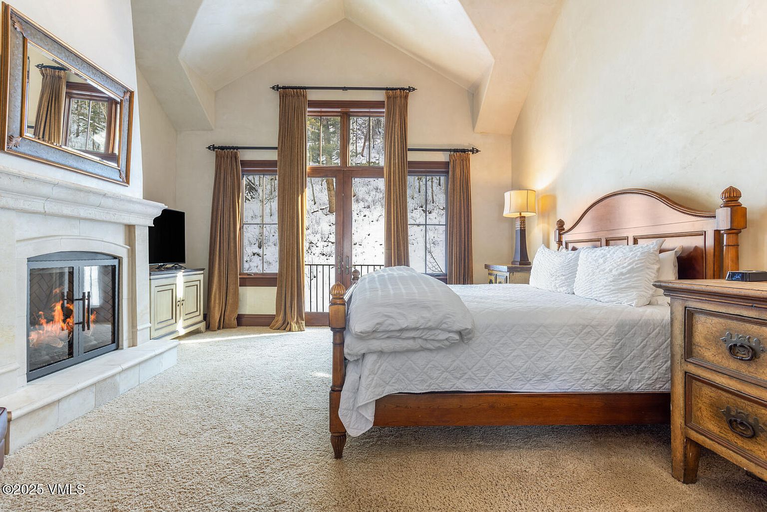 This is a primary bedroom featuring a fireplace, a large bed with white linens, and a view of a snowy landscape through a set of windows. The room has a warm and inviting feel, with neutral tones and natural light creating a cozy atmosphere. The architectural details, such as the vaulted ceiling and the stone fireplace, add to the room's character and appeal.
