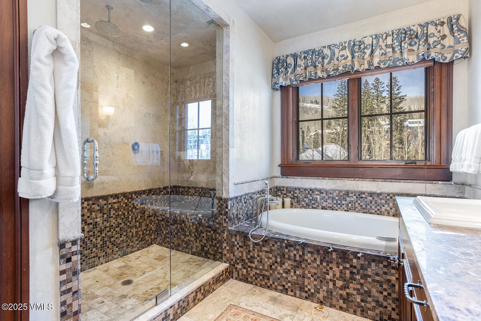 This is a primary bathroom featuring a glass-enclosed shower with a rainfall showerhead and a built-in bathtub with mosaic tile surround. A window with a valance provides natural light and a view of the outdoors. The bathroom has a luxurious and spa-like feel with neutral tones and high-end finishes.