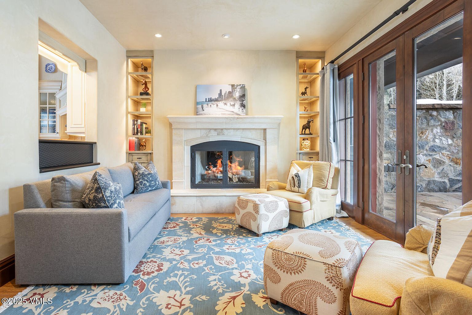 This inviting living room features a cozy fireplace flanked by built-in bookshelves, creating a warm and sophisticated atmosphere. A comfortable gray sofa and armchair with patterned ottomans provide ample seating, while a decorative rug adds a touch of color and elegance. French doors lead to an outdoor area, enhancing the room's appeal and natural light.