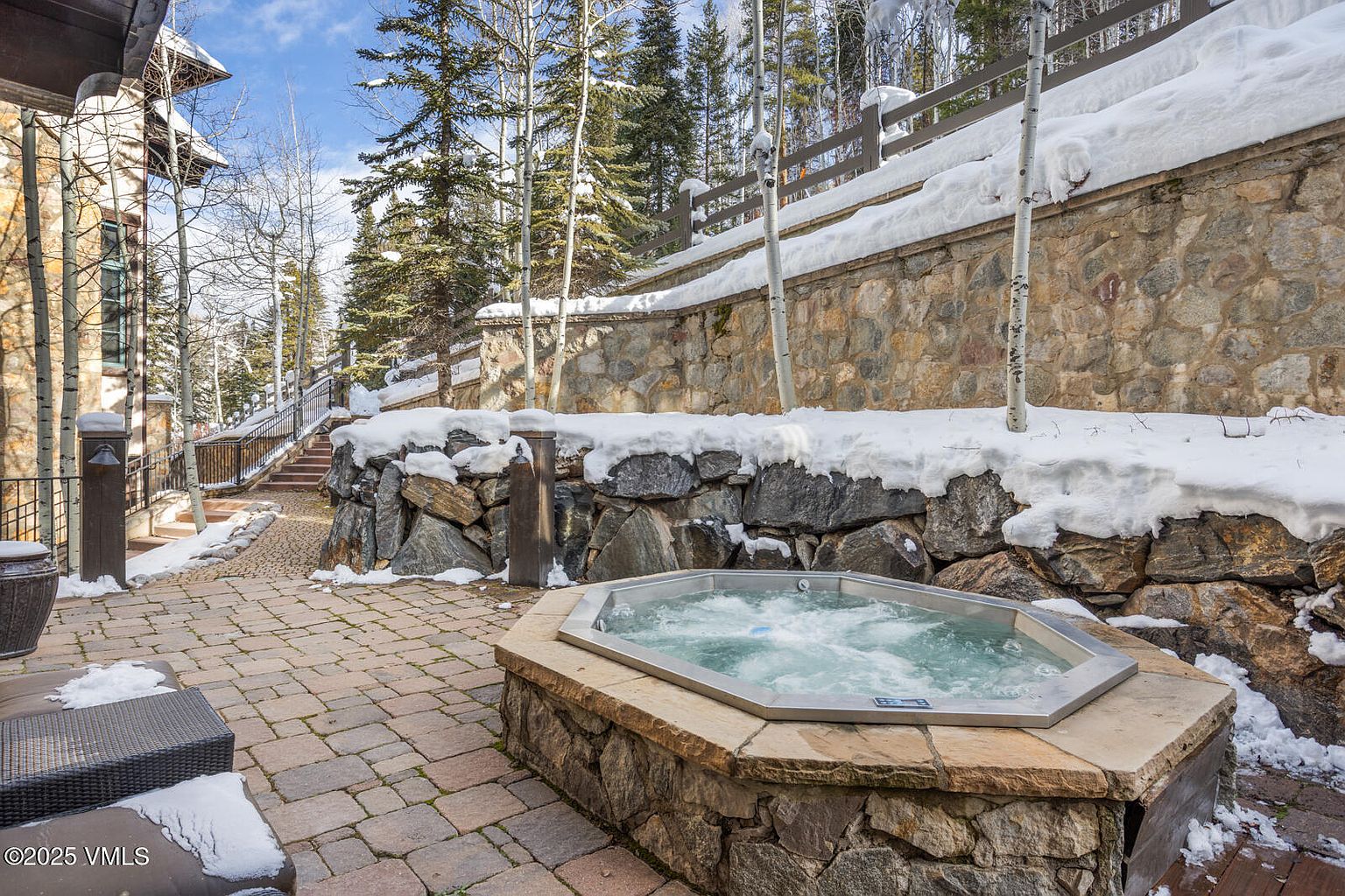 This image showcases an inviting outdoor patio area, complete with a stone-encased hot tub bubbling with water. The patio is paved with brick and features stone retaining walls, some covered in snow, adding to the mountain retreat ambiance. A glimpse of outdoor furniture suggests a comfortable space for relaxation and entertainment.