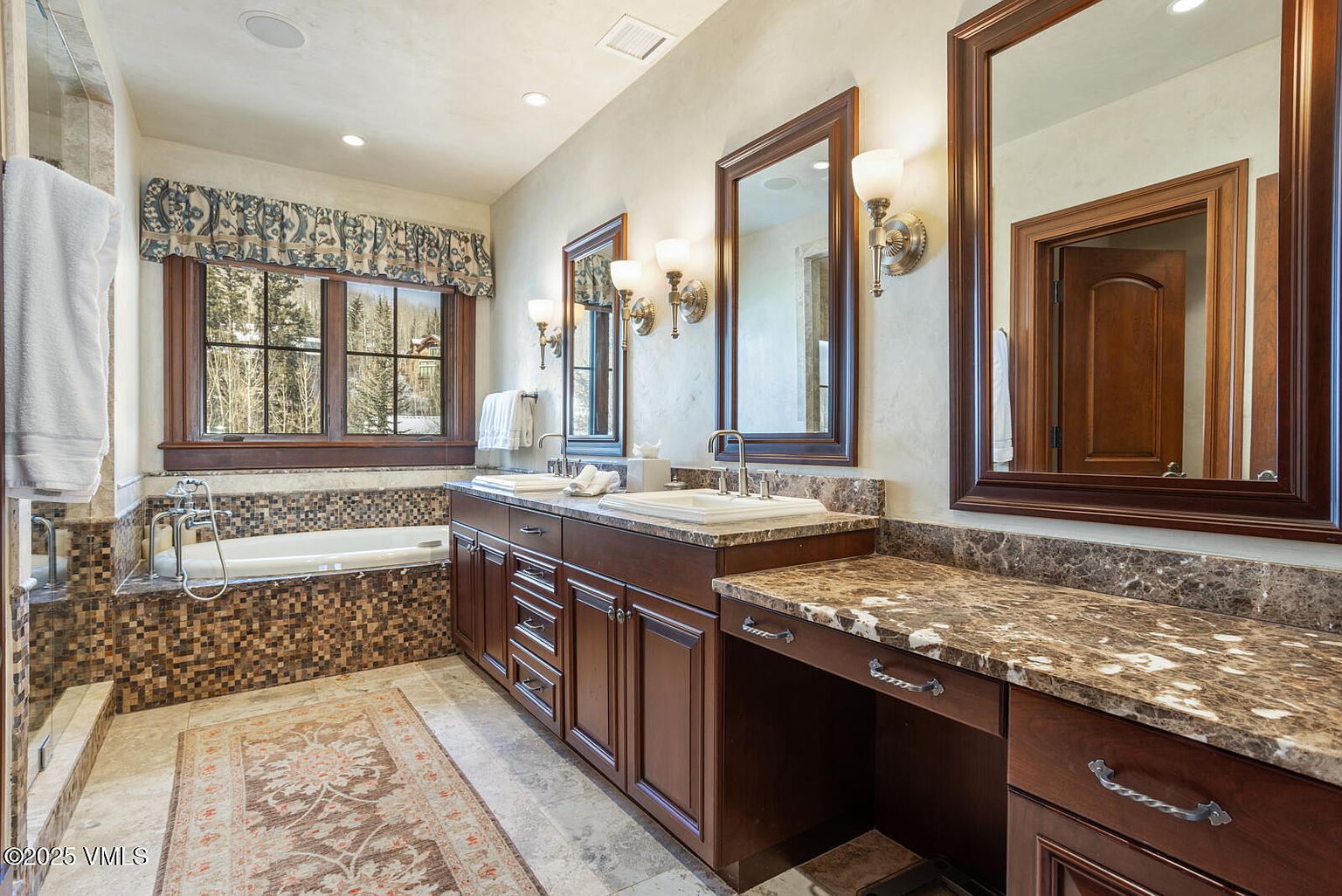 This is a luxurious primary bathroom featuring a double vanity with marble countertops and dark wood cabinetry. The room includes a soaking tub with mosaic tile surround and a large window with a patterned valance, providing natural light. Large framed mirrors and sconce lighting add to the elegant and well-appointed space.