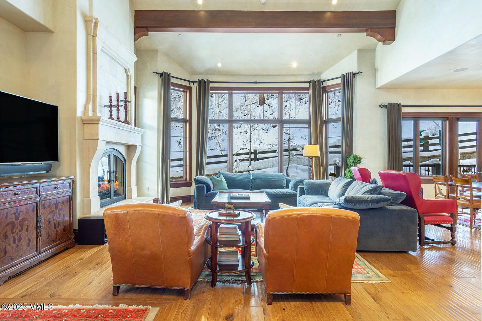 This is an interior shot of a living room featuring hardwood floors, a stone fireplace, and large windows offering a view of a snowy landscape. The room is furnished with comfortable sofas and armchairs, creating a cozy and inviting atmosphere. The perspective is from the entrance of the room, showcasing the entire living space.