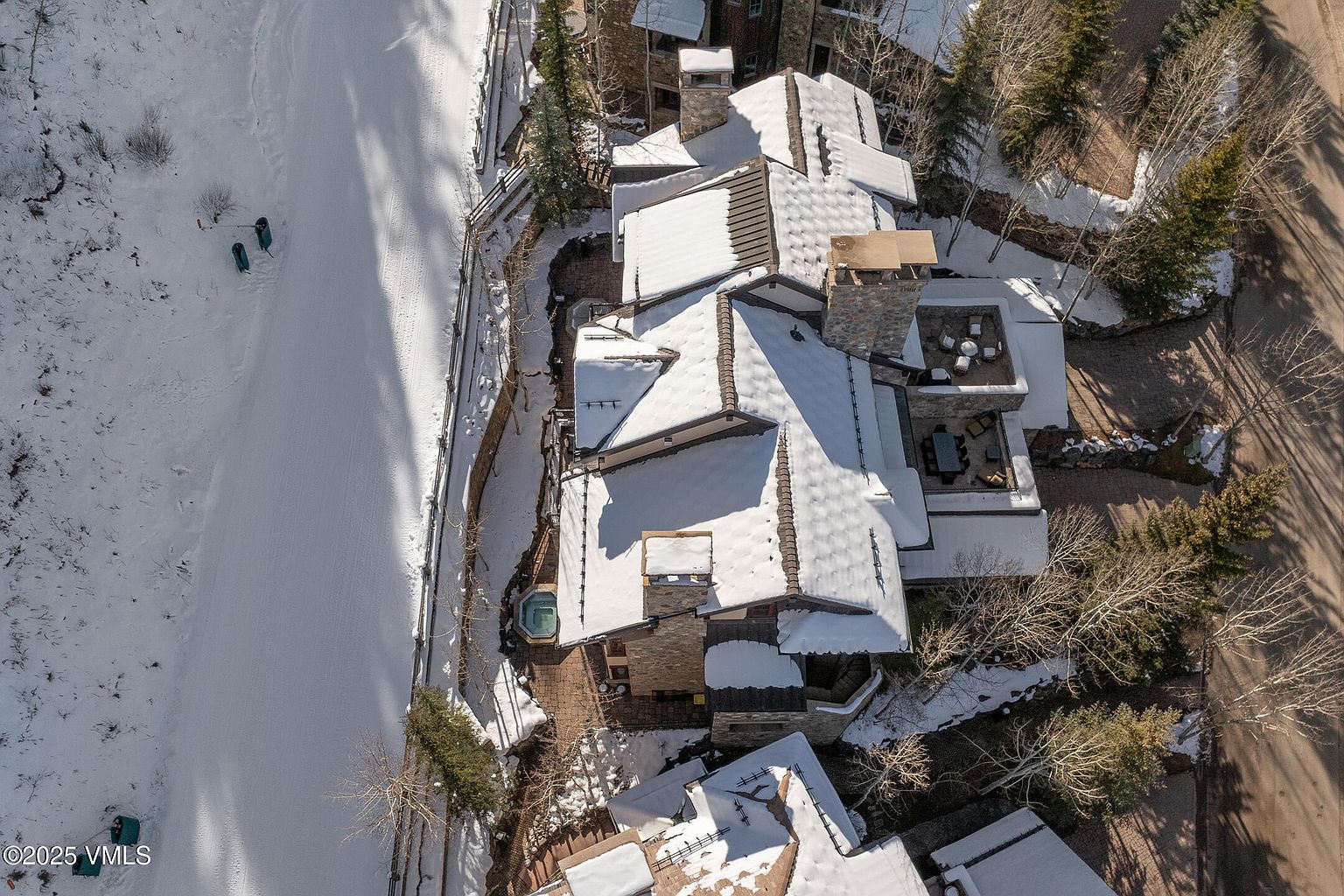 This aerial shot showcases a luxurious multi-story home with a complex roofline partially covered in snow. The property features stone and brick construction, multiple chimneys, and outdoor living spaces including a patio with furniture and a hot tub. The surrounding landscape includes snow-covered ground, bare trees, and a paved driveway, creating a serene winter scene.