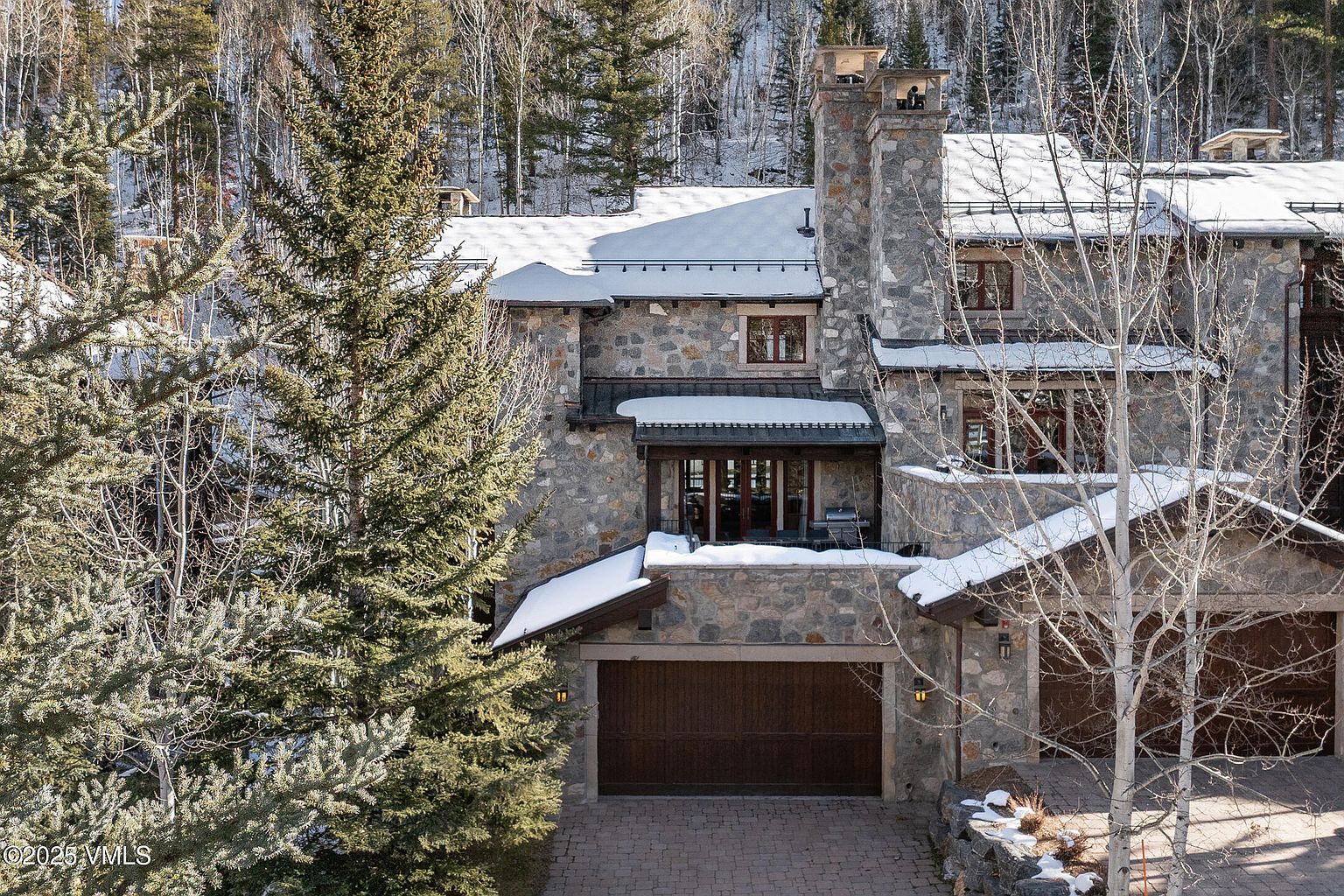 This is a front exterior view of a luxurious stone house with a multi-car garage. The house features a stone facade, a prominent stone chimney, and a roof partially covered in snow. Mature trees frame the house, adding to its secluded and upscale appeal.