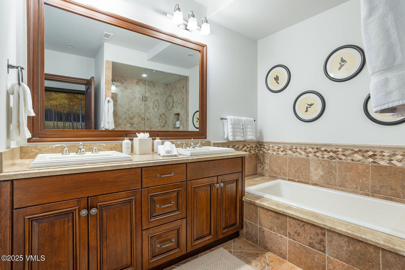 This is a primary bathroom featuring a double vanity with a wooden cabinet base and light-colored countertop. A large mirror hangs above the vanity, reflecting a tiled shower. The bathroom also includes a bathtub with tiled surround, and decorative framed artwork on the wall, creating a luxurious and well-appointed space.