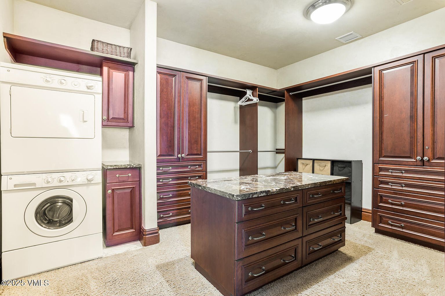 This is a well-organized walk-in closet featuring custom dark wood cabinetry and drawers, complemented by a central island with a marble countertop. A stacked washer and dryer unit is visible on the left, suggesting a combined laundry and closet space. The room is illuminated by a ceiling light, and the carpeted floor adds a touch of comfort.