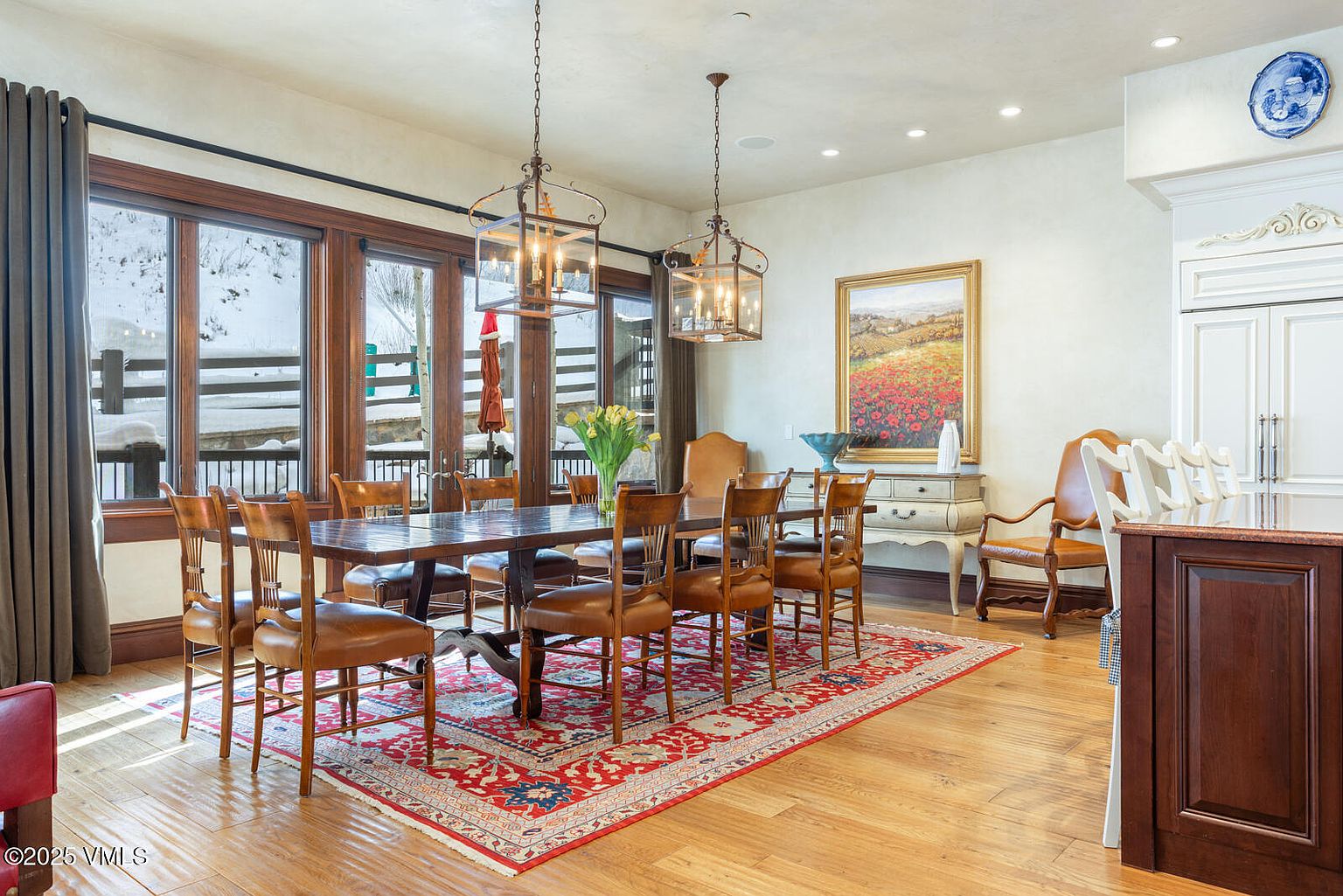This is an interior shot of a dining room featuring a large wooden dining table surrounded by leather chairs, set upon a red patterned rug. Natural light floods the room through large windows, complemented by two hanging lantern-style chandeliers. The room is decorated with a painting and a blue and white plate, creating a warm and inviting atmosphere.