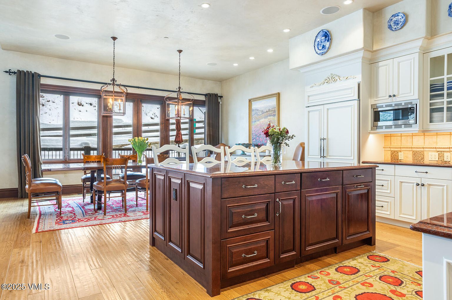 This is a well-lit kitchen featuring a large dark wood island with a light countertop, complemented by white cabinetry and stainless steel appliances. The kitchen also includes a dining area with a wooden table and chairs, illuminated by pendant lights, and a view of a snowy landscape through the windows. The overall impression is one of warmth and sophistication.