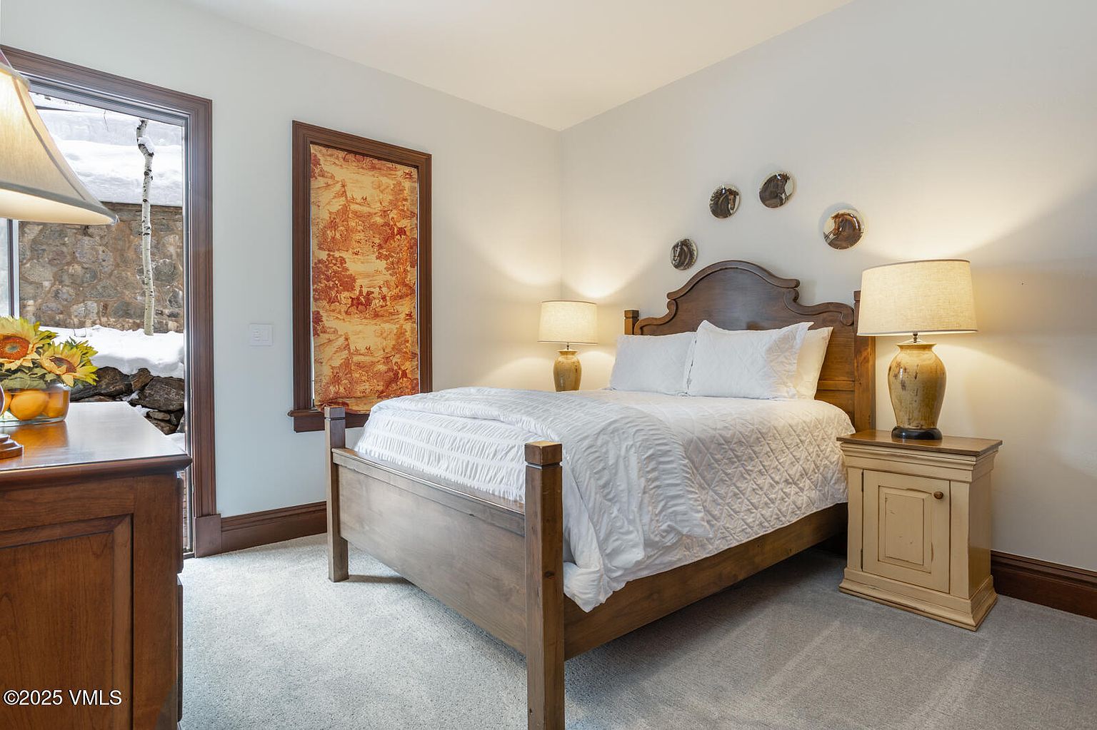This is a cozy guest bedroom featuring a wooden bed frame with white bedding, complemented by matching nightstands and lamps. A window with a patterned shade adds character, while decorative plates above the headboard enhance the room's charm. The overall impression is warm and inviting, perfect for a comfortable stay.