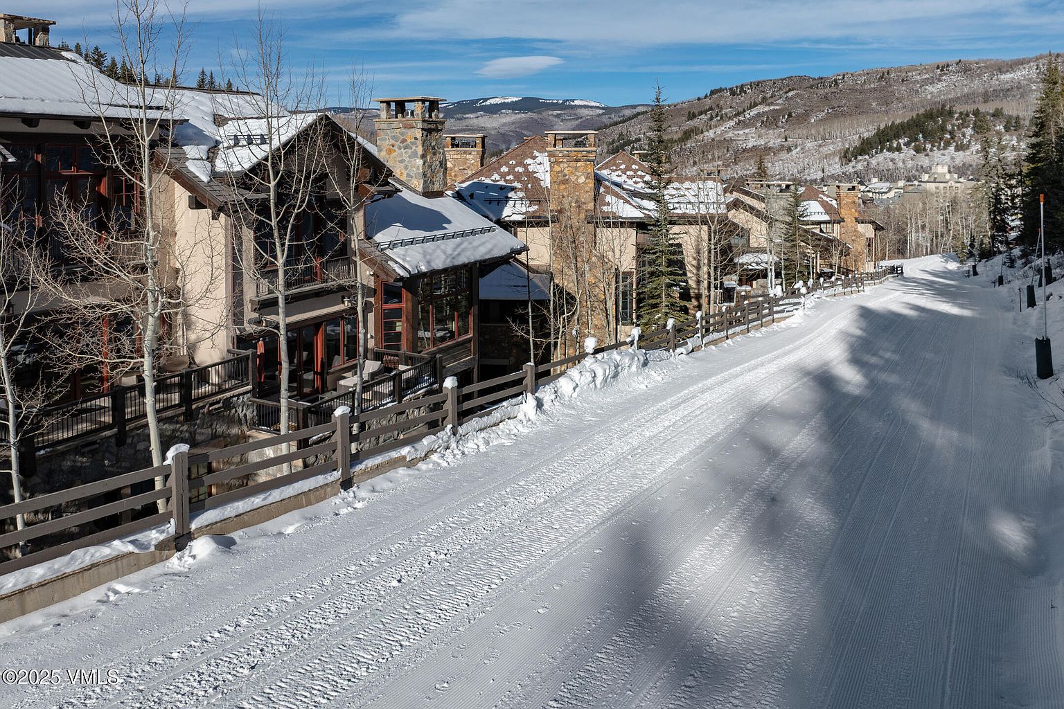 This image showcases a row of luxurious homes in a snowy, mountainous setting. The homes feature a mix of stone and stucco exteriors, with snow-covered roofs and stone chimneys. A snow-covered road runs alongside the properties, adding to the winter wonderland aesthetic. The scene evokes a sense of upscale mountain living and tranquility.