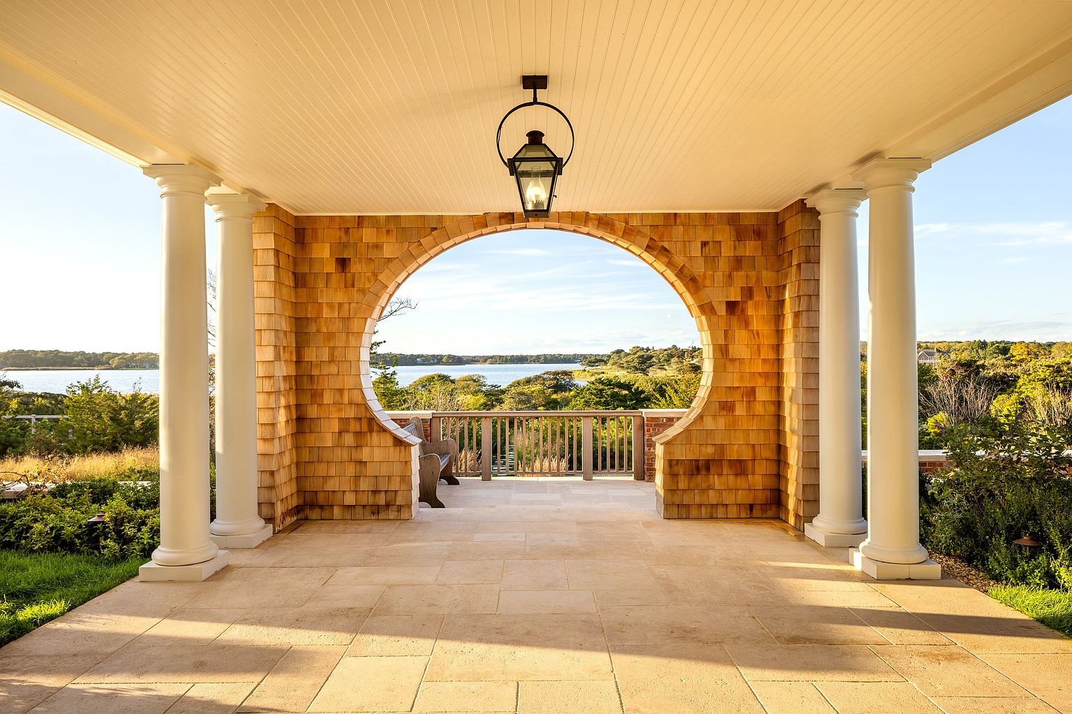 This elegant covered patio features a distinctive circular arched opening that frames a scenic view of the water and surrounding landscape. The space is defined by cedar shingle siding, classic white columns, and a stone-tiled floor, creating a sophisticated coastal aesthetic. A wooden bench sits near the railing, inviting relaxation while overlooking the tranquil outdoor setting.