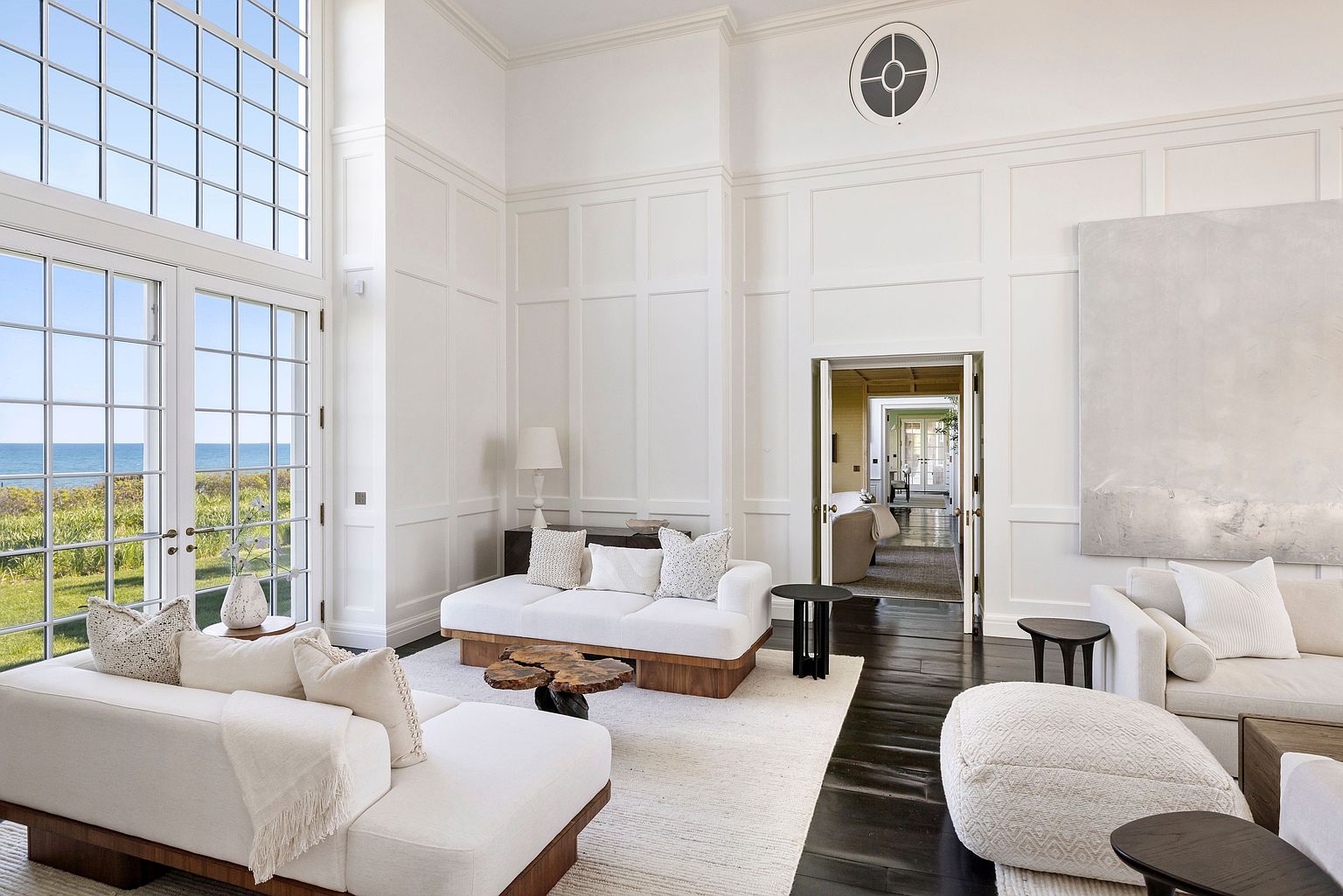 This expansive, light-filled living room features high ceilings with white paneling and dark wood flooring, creating a sophisticated and airy atmosphere. The space is furnished with plush, cream-colored sofas and a unique, organic-shaped wood coffee table, all centered on a large area rug. Large windows provide a stunning, unobstructed view of the ocean, while a doorway leads into a bright, connected hallway, emphasizing the home's open and flowing layout.