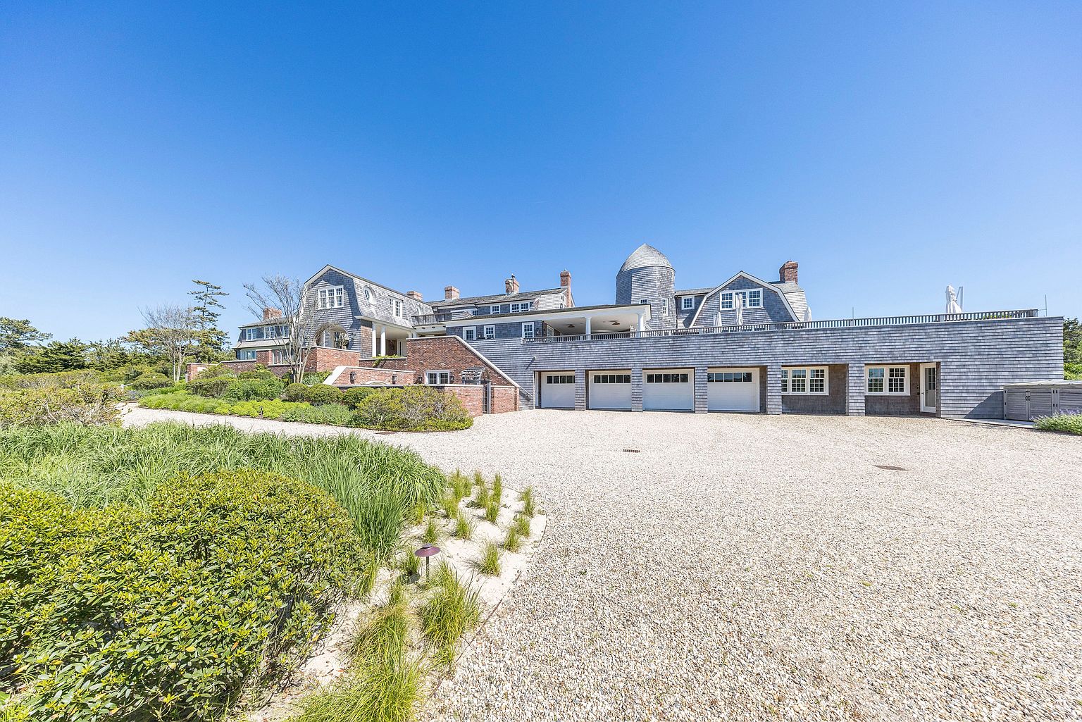 This expansive shingle-style estate features a multi-gabled roofline, a distinctive silo-inspired tower, and a long, integrated garage wing. The property is set against a clear blue sky, with a wide gravel driveway leading up to the residence, framed by manicured landscaping and native grasses. The architecture blends traditional coastal elements with a sprawling, modern layout, creating an impression of luxury and privacy.